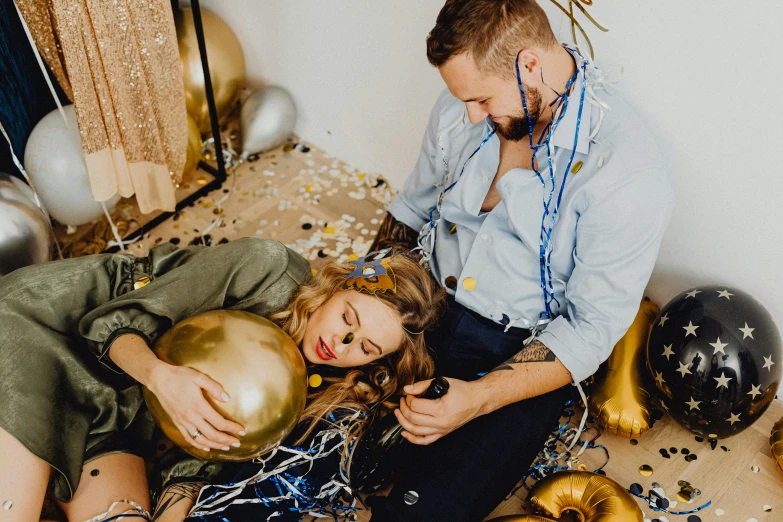 a man and woman sitting on the floor with balloons and confetti, by Julia Pishtar, trending on pexels, he holds her while she sleeps, gold theme, millaise and greg rutkowski, worn out