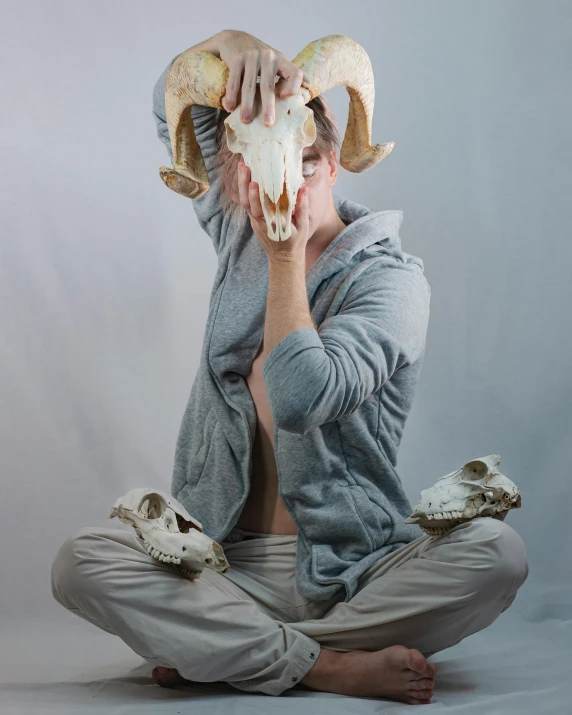 a man sitting on the ground with a goat skull on his head, inspired by Sarah Lucas, unsplash, non binary model, yoga pose, studio photo, ilustration