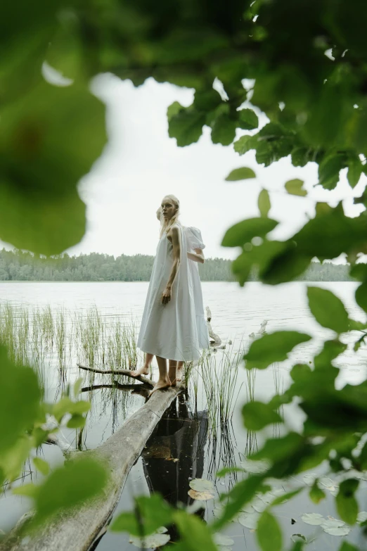 a woman standing on a log in the middle of a lake, by Grytė Pintukaitė, wearing a white folkdrakt dress, cottagecore, movie filmstill, with a white complexion