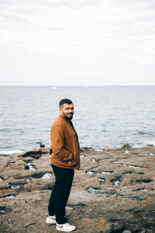 a man standing on top of a rock next to the ocean, he is wearing a brown sweater, a portrait of rahul kohli, manly, profile image