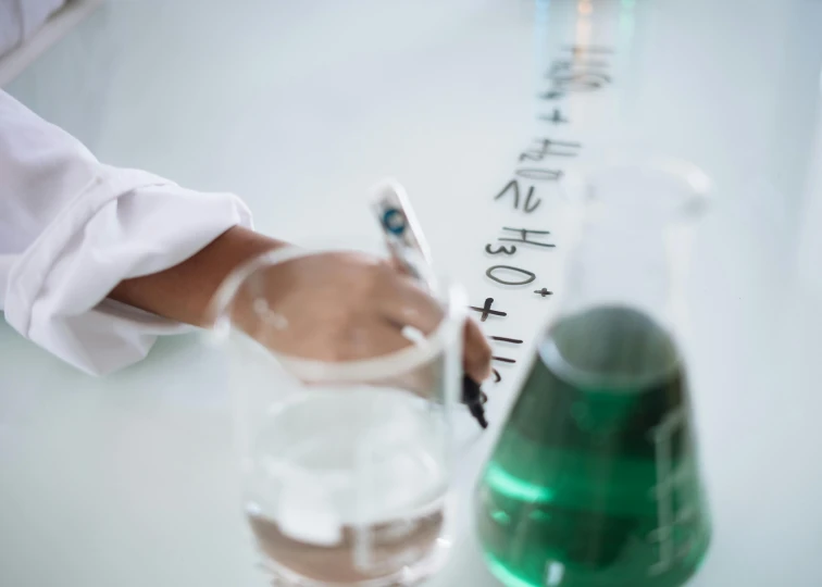 a close up of a person writing on a piece of paper, scientific glassware, green water, formulae, over-shoulder shot