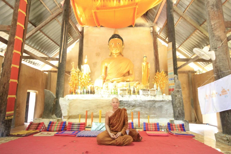 a man sitting in front of a large buddha statue, inside her temple, a blond, 🦩🪐🐞👩🏻🦳, monk clothes