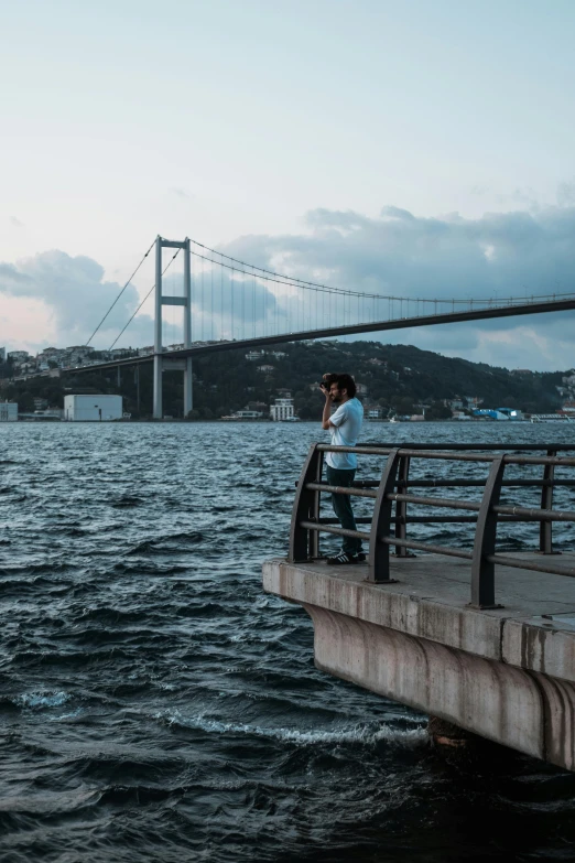 a man standing on top of a pier next to a body of water, by irakli nadar, tall bridge with city on top, istanbul, low quality photo, unsplash photography