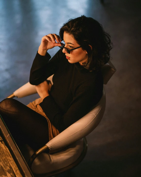 a woman sitting in a chair talking on a cell phone, a character portrait, inspired by Elsa Bleda, trending on unsplash, aestheticism, with square glasses, seductive stare, wearing a black sweater, emerging from her lamp