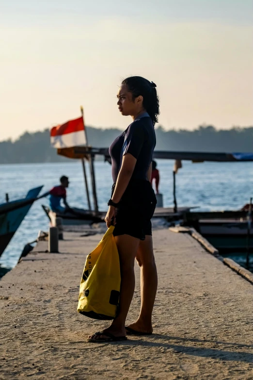 a woman standing on a pier next to a body of water, a picture, inspired by Steve McCurry, happening, indonesia, low sun, carrying survival gear, with yellow cloths