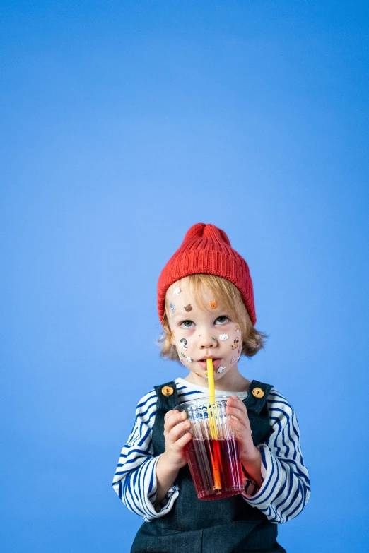 a little boy in a red hat drinking a drink, inspired by Hendrick Avercamp, pexels, pop art, red facial stripe, with a blue background, soda themed girl, press shot