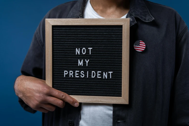 a man holding a sign that says not my president, by Julia Pishtar, trending on unsplash, stuckism, miscellaneous objects, avatar image, portrait n - 9, frank moth