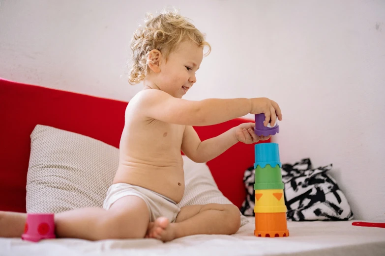 a baby sitting on a bed playing with a stack of blocks, by Dan Content, pexels contest winner, cups and balls, tower of babel, he is about 20 years old | short, multicolored