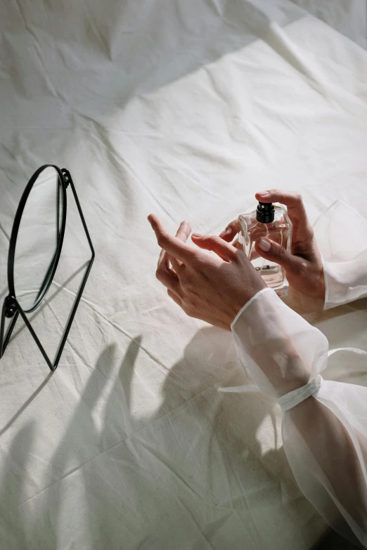 a woman laying on a bed holding a bottle of perfume, pexels contest winner, visual art, hands pressed together in bow, white clothing, sheer fabrics, issey miyake