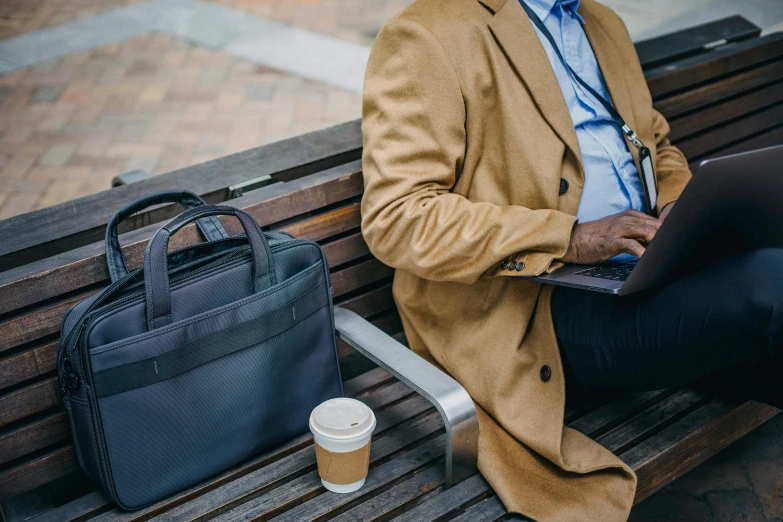 a man sitting on a bench with a laptop, by Carey Morris, holding a briefcase, celebration of coffee products, tailored clothing, trench coat and suit