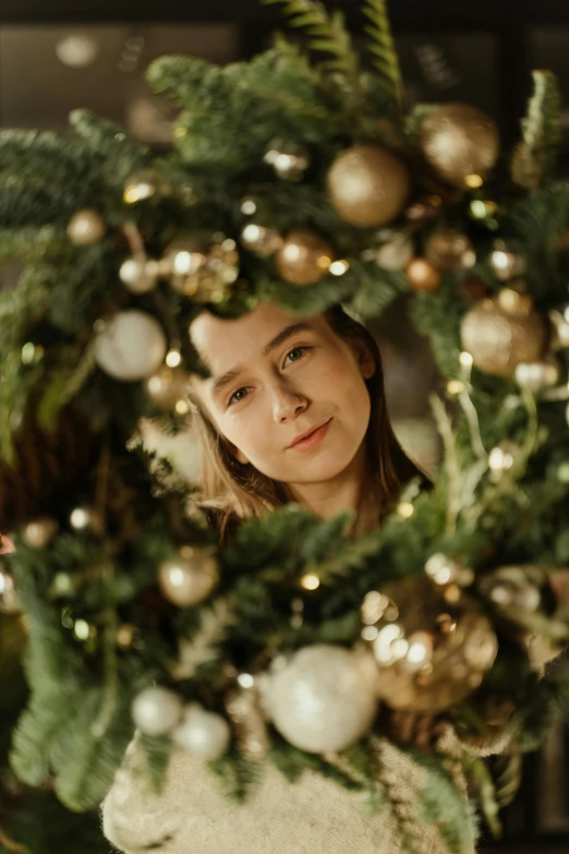 a woman standing in front of a christmas wreath, by Julia Pishtar, pexels contest winner, lovingly looking at camera, teen girl, golden ornaments, full frame image