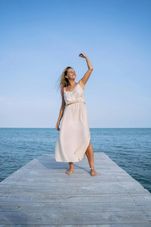 a woman in a white dress standing on a pier, pexels contest winner, happening, smiling and dancing, light tan, lighthearted, confident relaxed pose