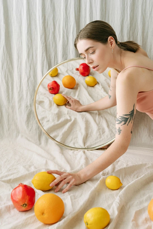 a woman sitting in front of a mirror surrounded by fruit, inspired by Kahlo, trending on pexels, arms of lemons, circular tattoo, laying on a bed, with a white background