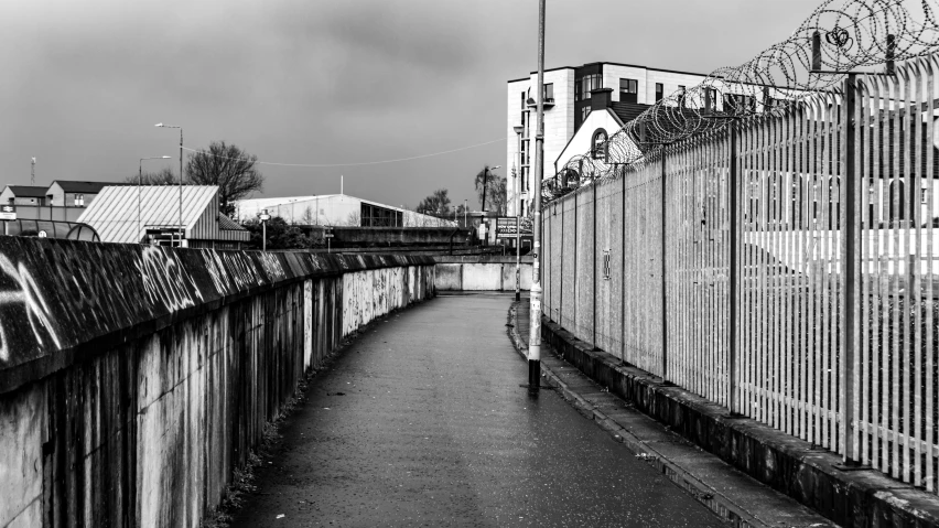 a black and white photo of a street next to a fence, inspired by Thomas Struth, unsplash, graffiti, wet aslphalt road after rain, paisley, waterway, empty metropolitan street