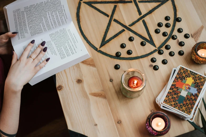 a person sitting at a table reading a book, by Julia Pishtar, trending on pexels, process art, pentacle, the candle is on a wooden table, witch clothes, pentagrams