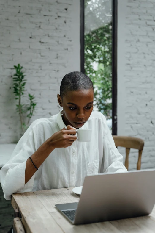 a woman sitting at a table with a laptop and a cup of coffee, wearing a white button up shirt, dark skinned, is ((drinking a cup of tea)), multiple stories