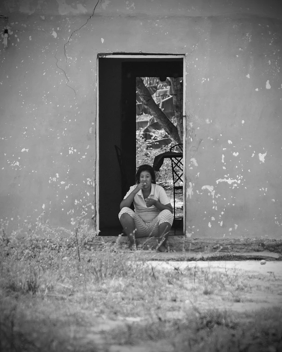 a black and white photo of a woman sitting in a doorway, inspired by Dorothea Lange, unsplash, in africa, an abandoned, outside on the ground, low colour