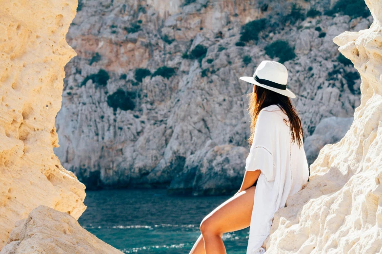 a woman sitting on a rock next to a body of water, tanned, holiday vibe, with hat, white clothes