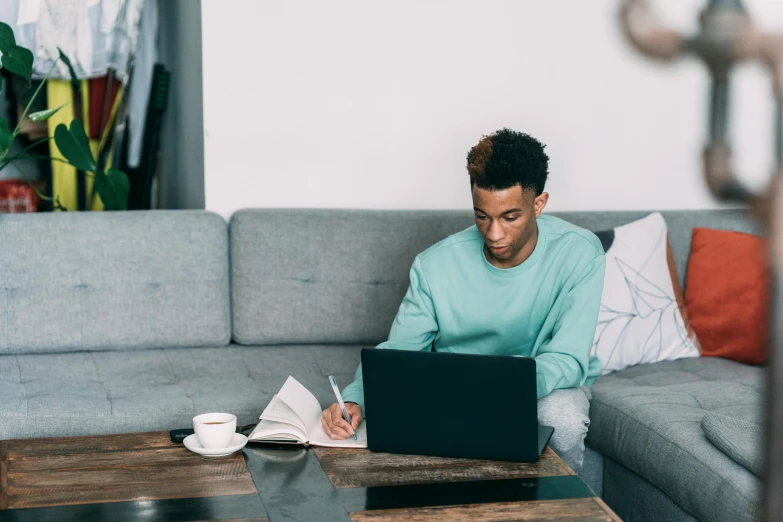 a man sitting on a couch working on a laptop, by Carey Morris, trending on pexels, happening, black teenage boy, pen and paper, teal aesthetic, table in front with a cup