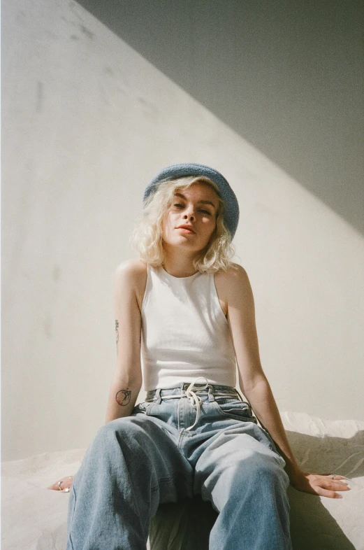 a woman sitting on top of a bed wearing a hat, inspired by Elsa Bleda, trending on pexels, photorealism, short curly blonde haired girl, baggy jeans, billie eilish portrait, soft cool colors