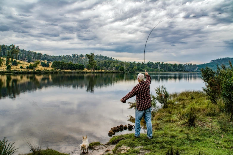 a man standing on top of a lush green field next to a lake, fishing pole, lachlan bailey, action photograph, wide river and lake