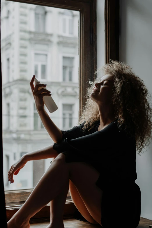 a woman sitting on a window sill in front of a window, a picture, trending on pexels, renaissance, hair fluid, mirror selfie, curly blond, holding a bottle