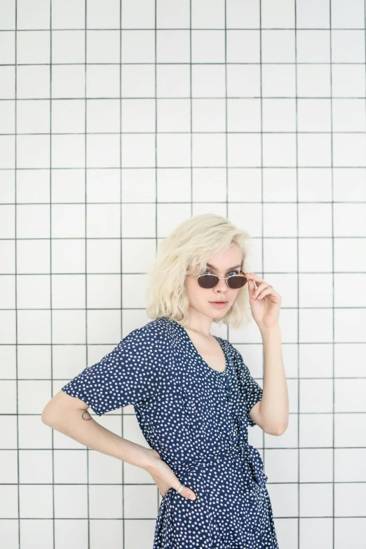 a woman standing in front of a tiled wall, inspired by Elsa Bleda, trending on unsplash, wearing blue sunglasses, extremely pale blond hair, drawn with dots, outfit: cop