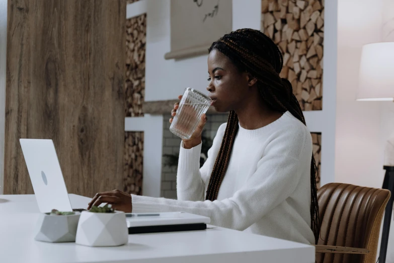 a woman sitting at a table with a laptop drinking a glass of water, with brown skin, curated collections, facing sideways, chilly