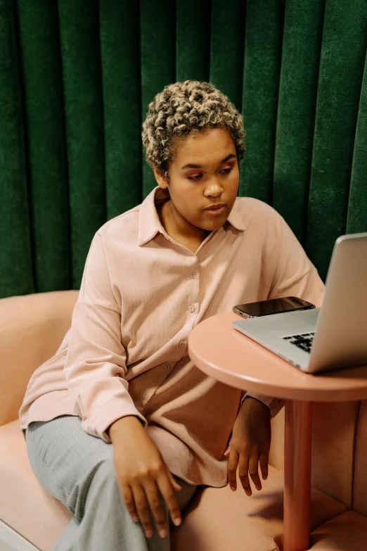 a man sitting on a couch using a laptop, by Carey Morris, pexels, happening, short blonde afro, wearing a light - pink suit, infp young woman, non-binary
