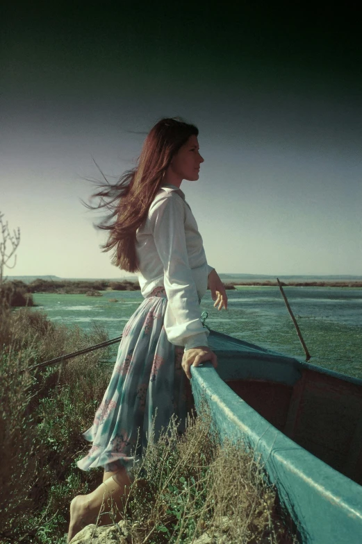 a woman standing next to a blue boat on the shore, an album cover, by Oleg Oprisco, renaissance, in the steppe, 1960s color photograph, white shirt and green skirt, liv tyler