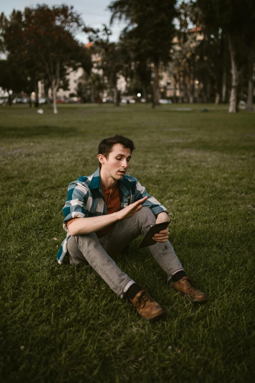 a man sitting on top of a lush green field, an album cover, by Ryan Pancoast, unsplash contest winner, realism, wearing a plaid shirt, in a city park, androgynous male, tinder profile