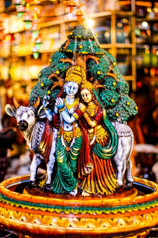 a statue sitting on top of a table next to a cow, by Shekhar Gurera, pexels contest winner, cloisonnism, 3 nymphs circling a fountain, festive colors, tree's, market