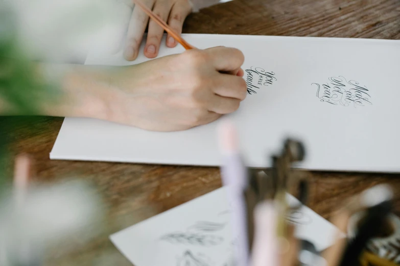 a person sitting at a table writing on a piece of paper, a drawing, by Emma Andijewska, trending on pexels, estrange calligraphy, costume design made with love, banner, crafts