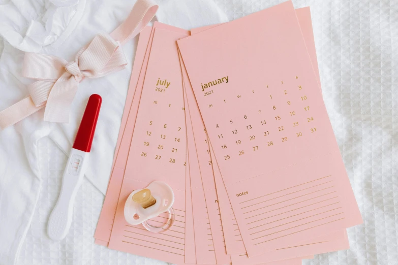 a pink calendar sitting on top of a bed next to a cup of coffee, by Julia Pishtar, trending on pexels, standing in gold foil, cards, thumbnail, skincare