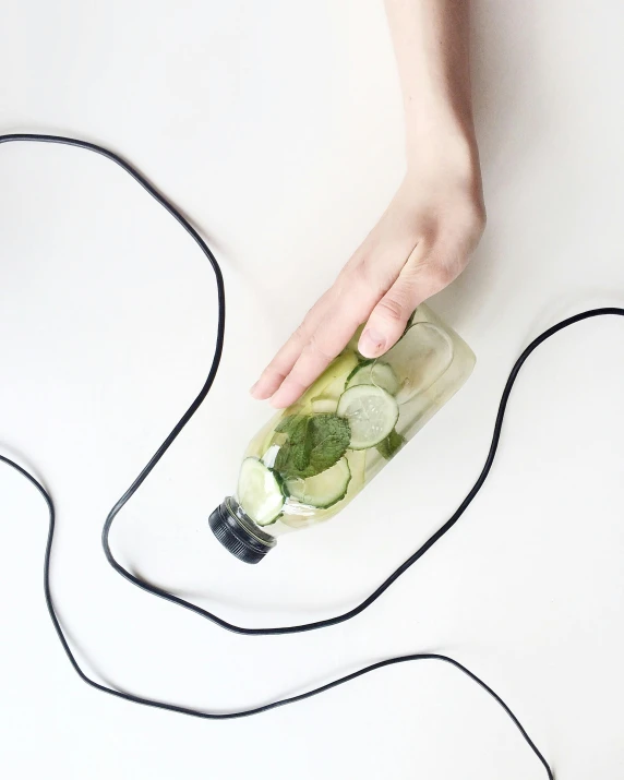 a person holding a bottle with cucumbers in it, by Nina Hamnett, happening, earphones, black wired cables, silicone skin, lemonade