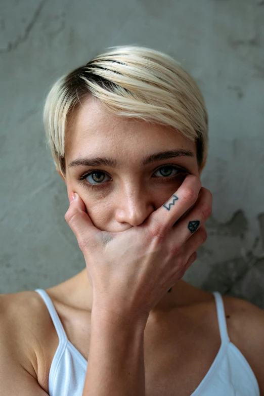 a woman covering her face with her hands, a tattoo, trending on pexels, antipodeans, blonde hair and large eyes, non binary model, hand over mouth, pixie cut