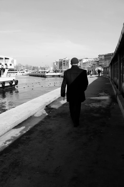 a man walking down a sidewalk next to a body of water, a black and white photo, istanbul, man in black suit, city docks, composer