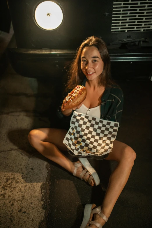 a woman sitting on the ground eating a donut, by Drew Tucker, pexels contest winner, photorealism, checkered floor, nightlife, hot dog, in front of a garage