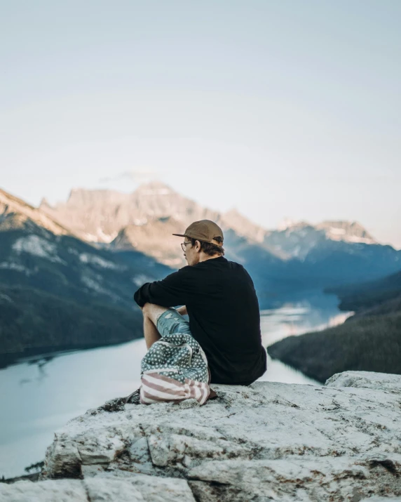a man sitting on a rock overlooking a lake and mountains, by Emma Andijewska, he also wears a grey beanie, towering above a small person, profile image, conde nast traveler photo