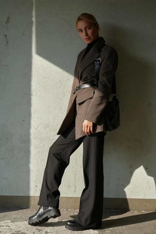 a woman standing in front of a white wall, by Nina Hamnett, wearing a worn out brown suit, yohji yamamoto, afternoon sunlight, wearing urban techwear