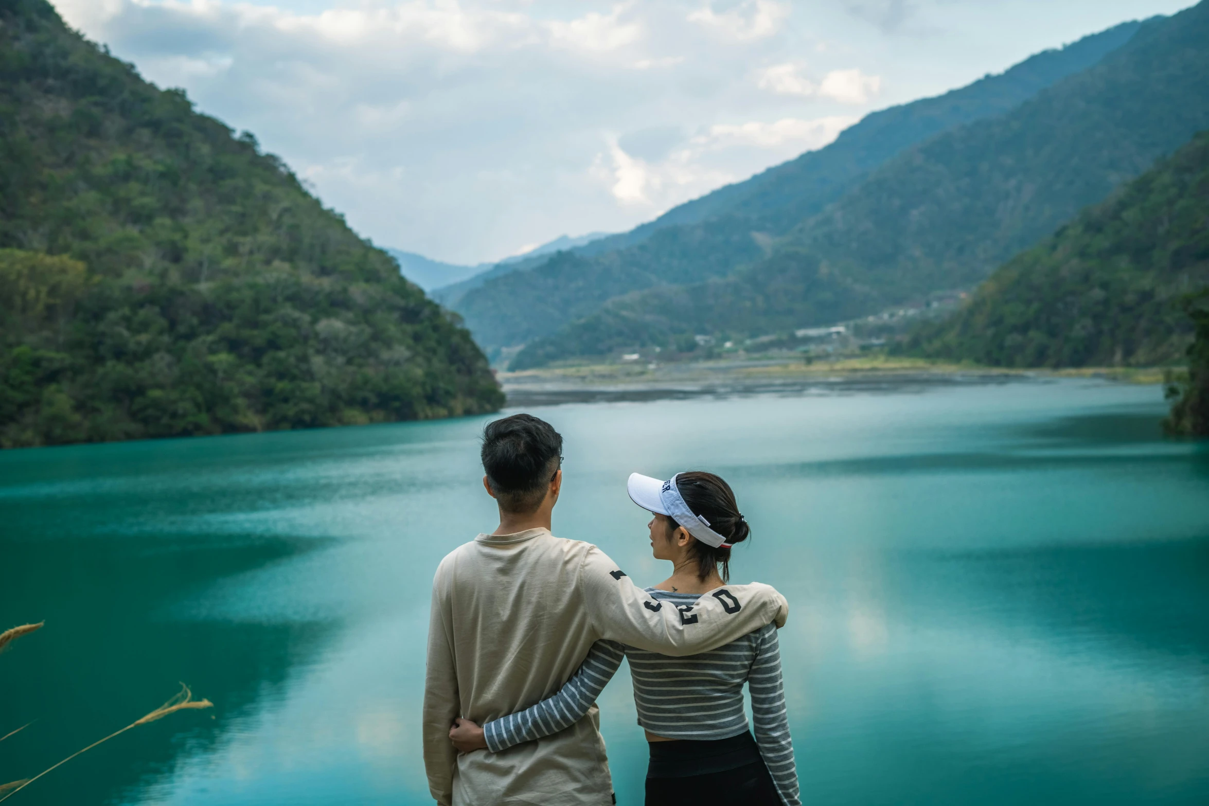 a couple standing next to each other in front of a lake, by Simon Gaon, pexels contest winner, hurufiyya, taiwan, blue and green water, overlooking a valley, outlive streetwear collection