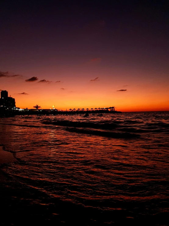 a couple of people standing on top of a beach next to the ocean, during the night, orange skies, miami beach, red skies