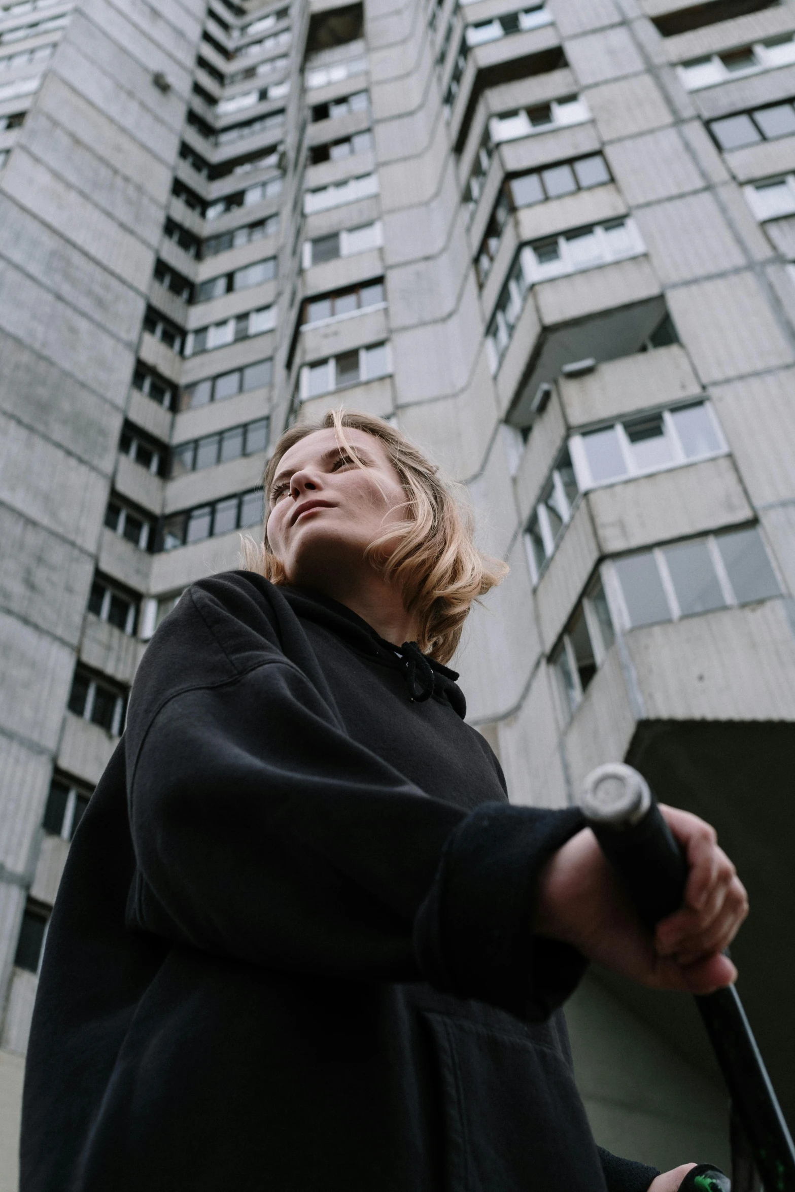 a woman standing in front of a tall building, by Christen Dalsgaard, unsplash, brutalism, wearing jeans and a black hoodie, holding a knife, portrait of homelander, kirsi salonen