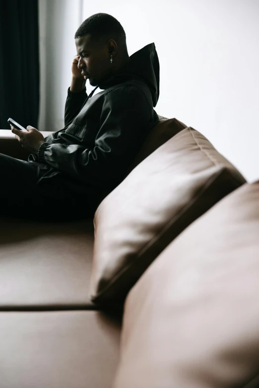 a man sitting on a couch talking on a cell phone, by Sebastian Spreng, trending on pexels, in a black hoodie, faceless people, leather sofa, androgynous person