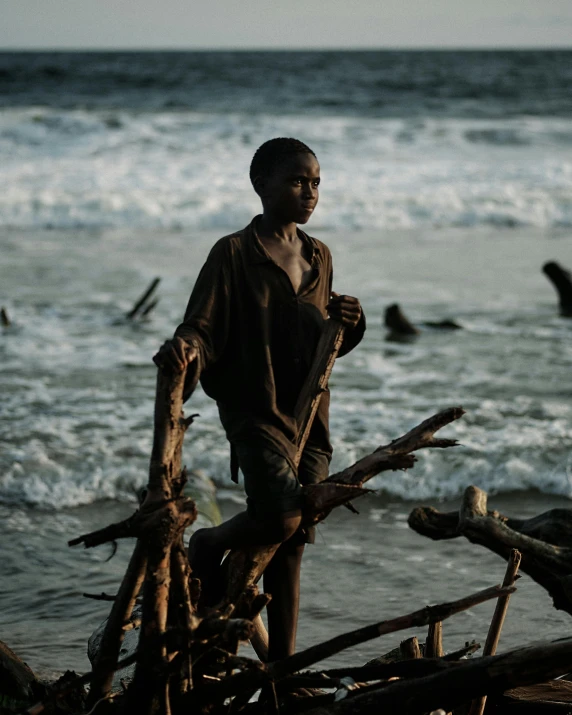 a man standing on top of a beach next to the ocean, an album cover, pexels contest winner, sumatraism, adut akech, cinematic movie still, young boy, driftwood sculpture