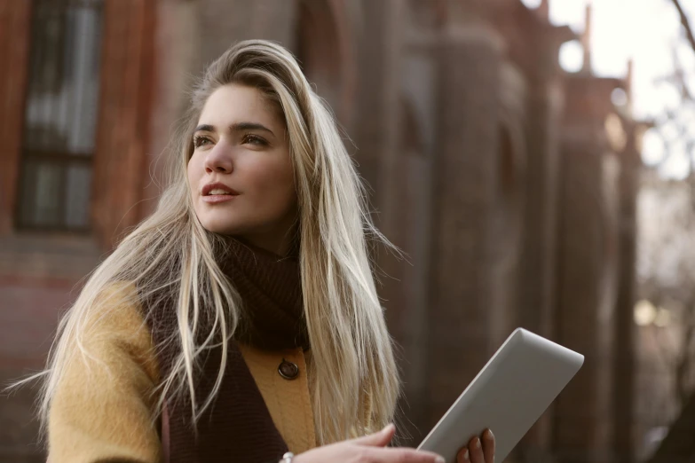 a woman holding a tablet computer in front of a building, a portrait, by Alice Mason, trending on pexels, beautiful blonde girl, brown, autum, looking upwards