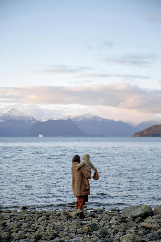 a person standing on a rocky beach next to a body of water, with a kid, mountains and oceans, private moment, snowy fjord