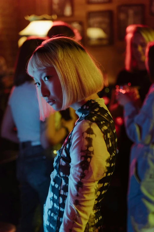 a woman standing in front of a group of people, inspired by Nan Goldin, trending on pexels, realism, 8 0 s asian neon movie still, with a bob cut, a girl with blonde hair, patterned