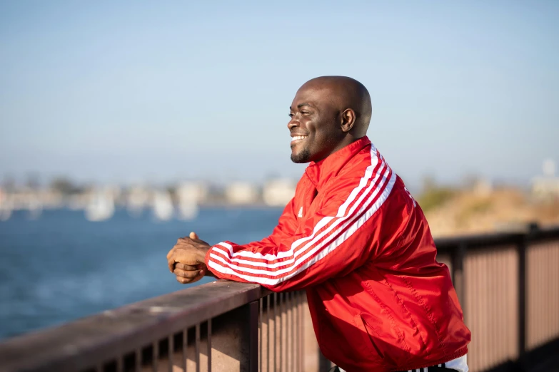 a man leaning on a railing next to a body of water, a portrait, by Stokely Webster, happening, wearing a track suit, red cloth around his shoulders, emmanuel shiru, oceanside