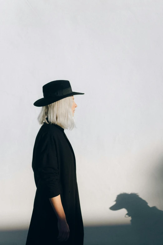 a woman standing in front of a shadow of a dog, inspired by Louisa Matthíasdóttir, trending on unsplash, minimalism, grey hair and a cowboy hat, wearing black modern clothes, profile image, long length slick white hair